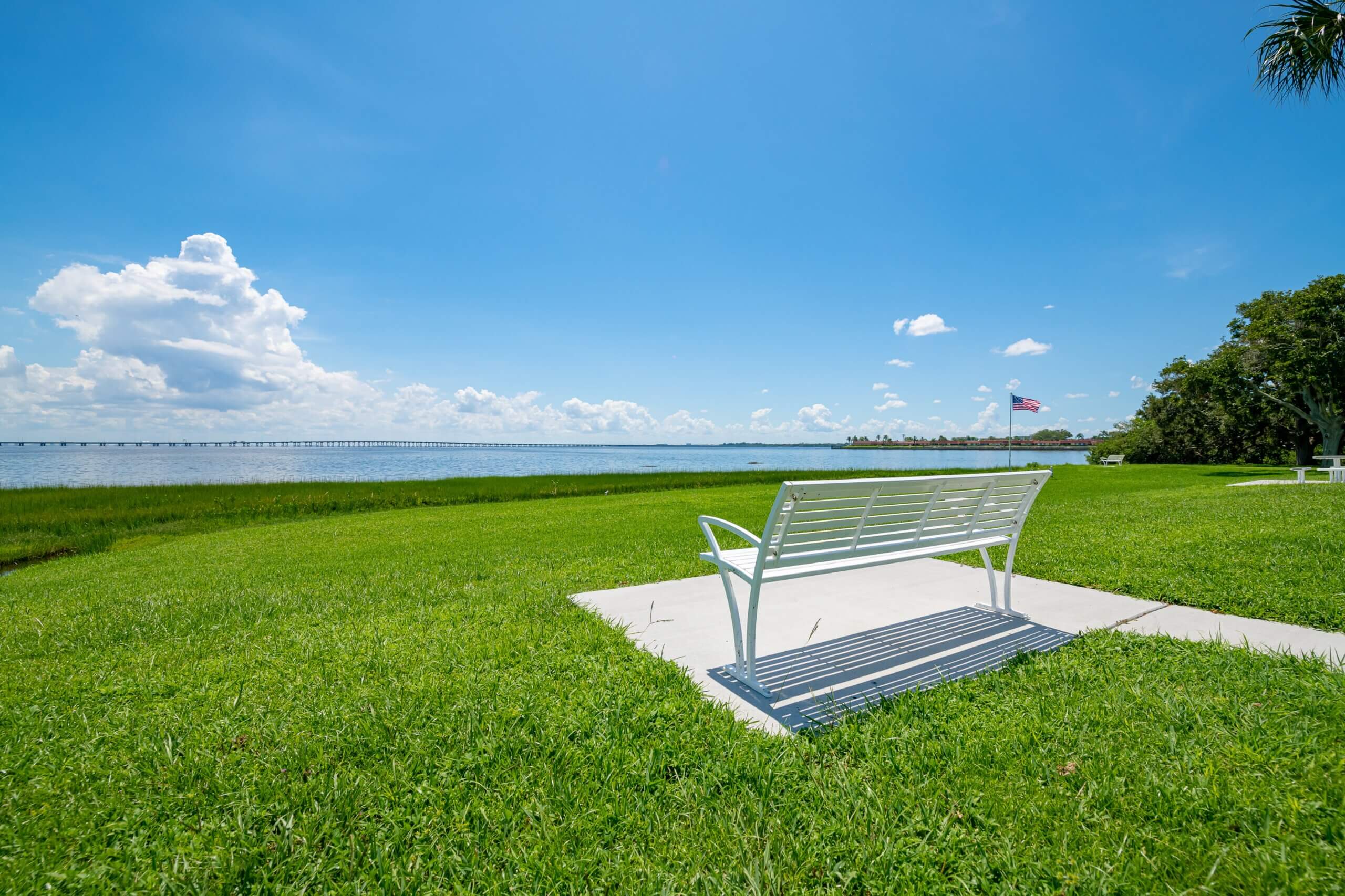 Outdoor white bench on the grass, facing the St. Pete Florida bay under a clear sky at Bayside Waters