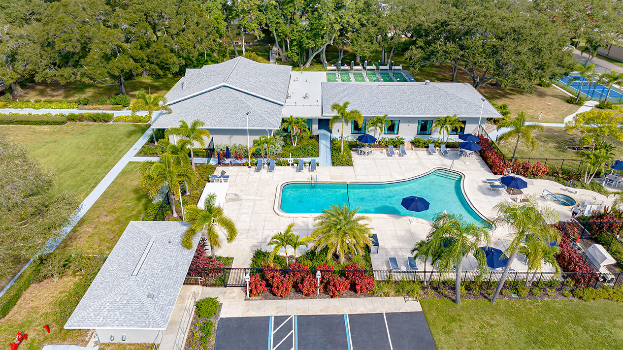 Aerial view of community pool area with loungers and umbrellas on a sunny day, adjacent to a clubhouse.