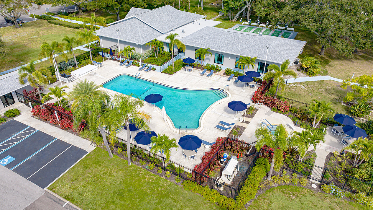 Aerial view of a community pool with blue umbrellas, surrounded by palm trees and adjacent to a clubhouse.