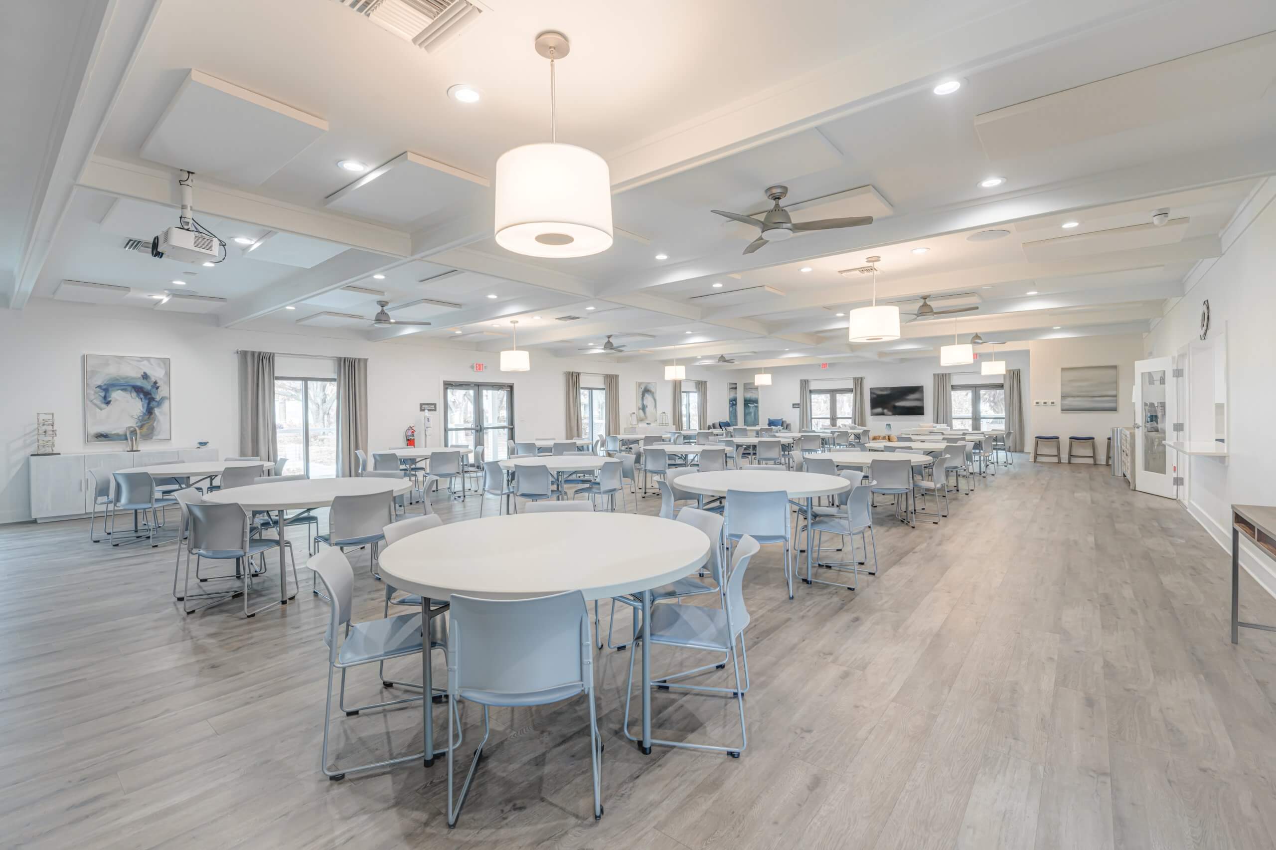 An expansive, modern dining and meeting hall with multiple round tables and chairs, large windows, and abstract wall art, complemented by high ceilings with stylish light fixtures.