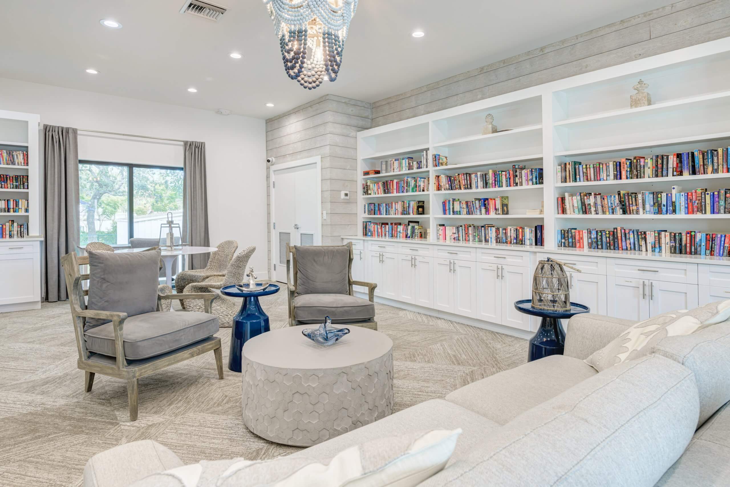 Bright living room with a gray sectional couch, two plush armchairs, a white built-in bookshelf filled with books, and a chandelier.