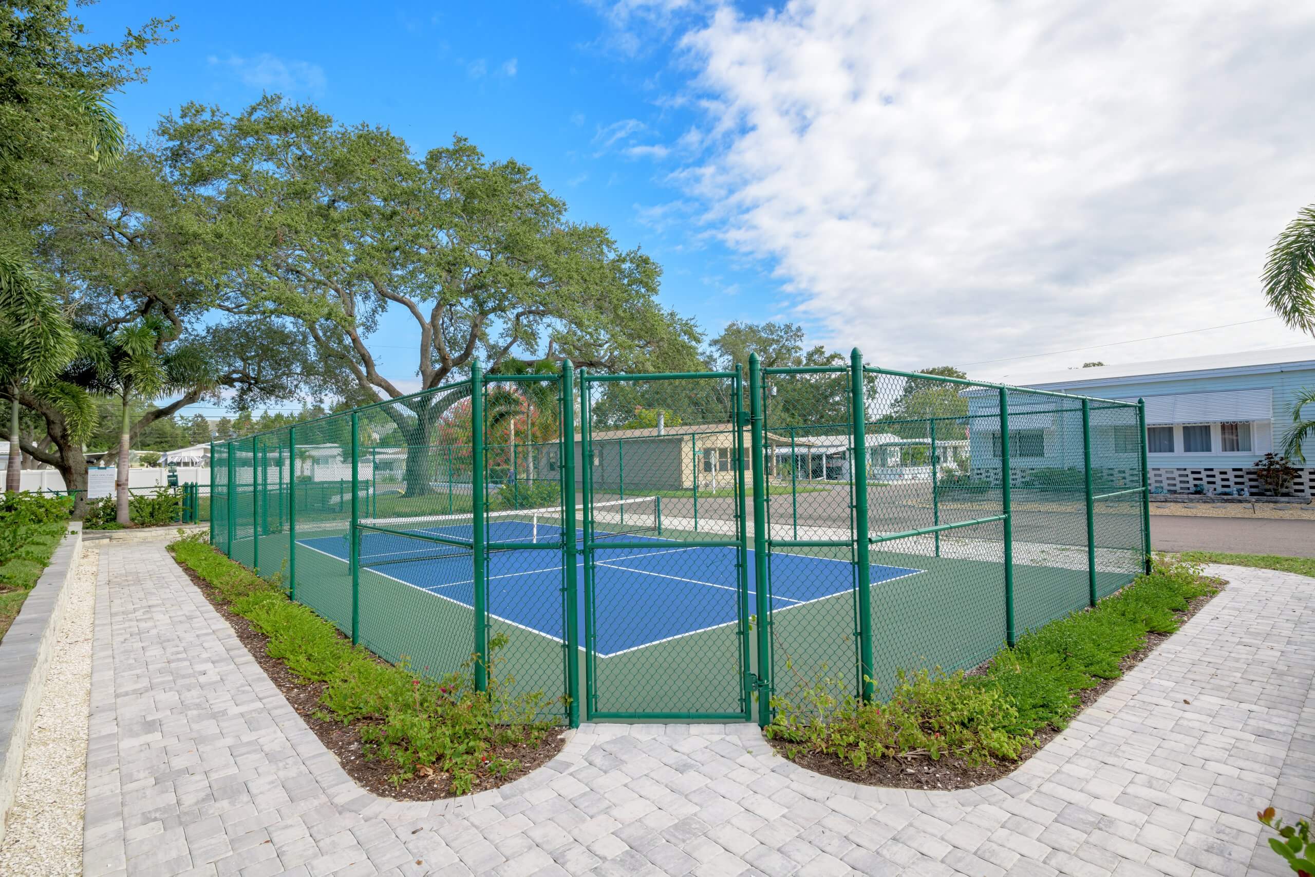 Community outdoor pickleball court enclosed by a green fence with surrounding trees and cobblestone pathways.